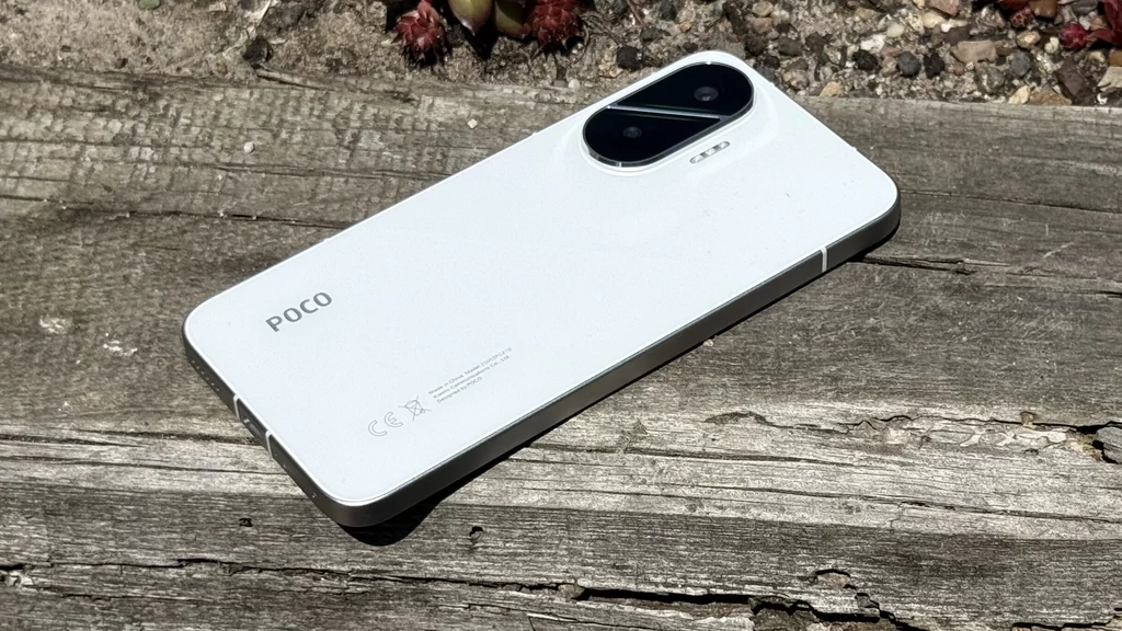 Um smartphone POCO F7 prateado, com o logo da POCO visível na parte inferior, está sobre uma superfície preta texturizada. A câmera traseira dupla é proeminente na parte superior esquerda.