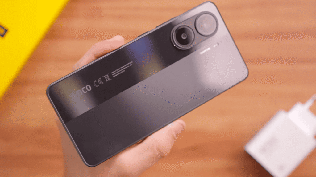 A imagem apresenta uma mão segurando o smartphone Poco X7 Pro na cor preta. A câmera dupla traseira é visível na parte superior do aparelho. Abaixo, a marca "POCO" e outras informações regulatórias estão impressas na tampa traseira. Ao lado do smartphone, sobre uma superfície de madeira clara, encontra-se um carregador branco. No canto superior esquerdo, desfocada, aparece uma parte de uma caixa amarela, possivelmente a embalagem do celular. A mão que segura o dispositivo está bem iluminada, assim como o smartphone, enquanto o fundo permanece suavemente desfocado, direcionando o foco para o Poco X7 Pro e seus acessórios próximos.