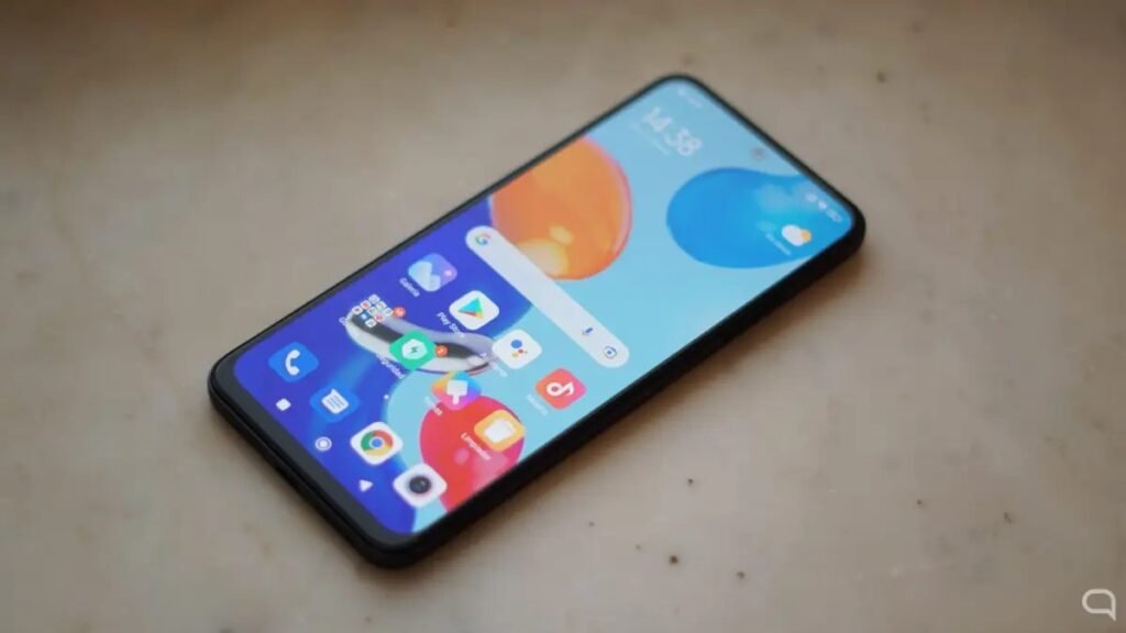 Um smartphone Xiaomi com a tela inicial ligada, mostrando o que parece ser a interface do Xiaomi HyperOS 3. A tela de início exibe ícones de aplicativos, a barra de pesquisa do Google, widgets e um papel de parede com formas abstratas em laranja e azul. O aparelho está apoiado sobre uma superfície clara e texturizada.