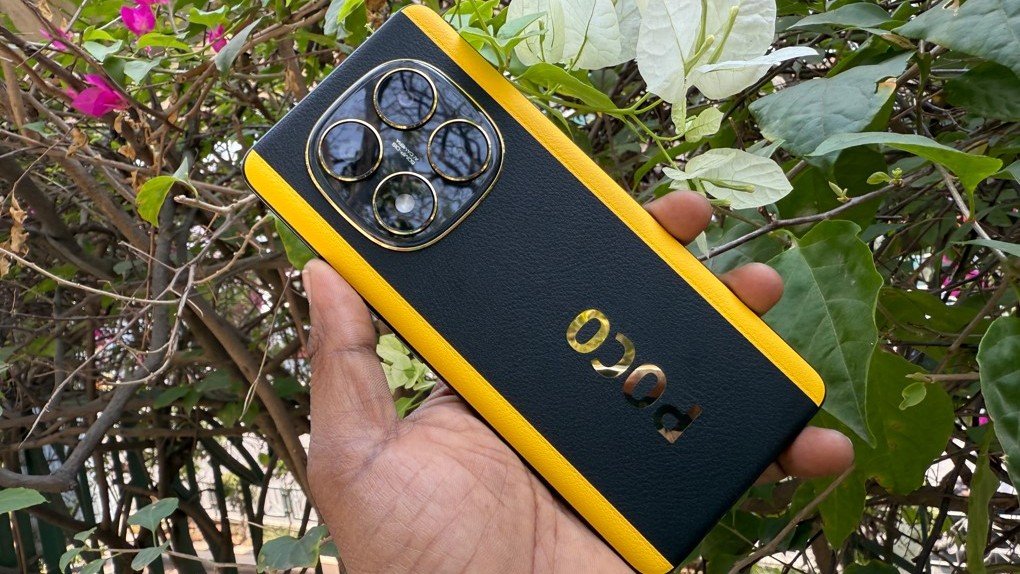Uma mão segura o smartphone POCO X7 preto com detalhes em amarelo dourado, destacando o módulo de quatro câmeras, em um ambiente externo com vegetação.