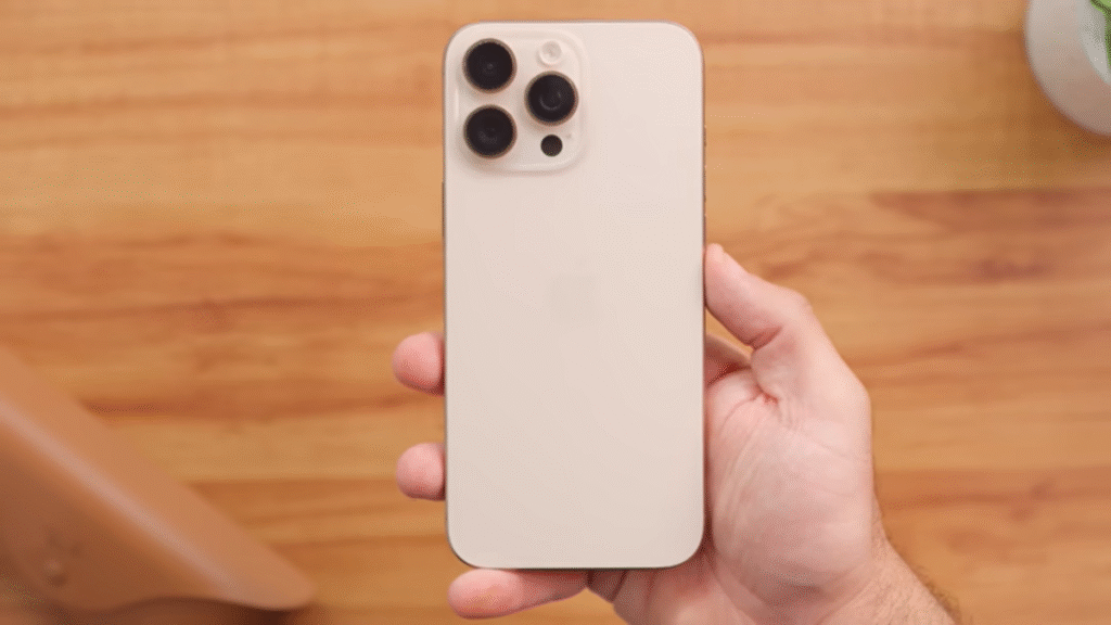 A imagem apresenta a mão de uma pessoa segurando firmemente um iPhone 16 Pro Max de cor clara, com a parte traseira do dispositivo voltada para a câmera. O destaque é o módulo de câmera tripla, com lentes proeminentes e um flash. A maçã, logotipo da Apple, está sutilmente visível no centro da traseira. À esquerda, parcialmente visível e fora de foco, encontra-se um smartphone preto, que se presume ser o Poco X7 Pro, sugerindo uma comparação entre os dois modelos. O fundo é composto por uma superfície de madeira clara, com um objeto bege à esquerda e um vaso branco com uma planta à direita, também parcialmente desfocados. A iluminação é suave e uniforme, realçando o design do iPhone.